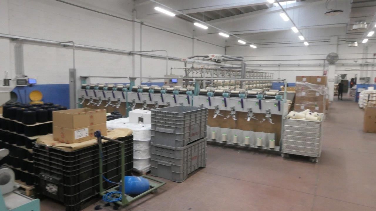 Il pavimento chiaro di un magazzino, con macchinari tessili industriali, pile di casse di plastica e scatole per spedizioni.
