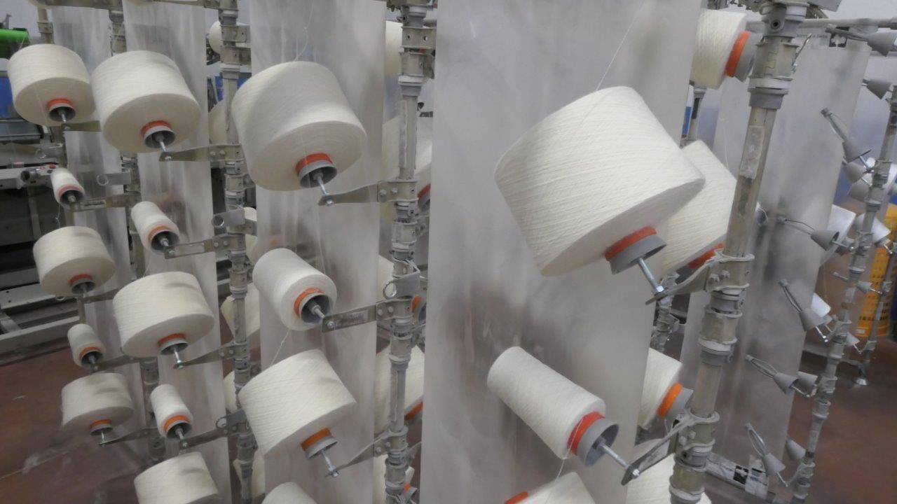 File di coni di filato bianco montati su un telaio porta-tessuti in un ambiente industriale.