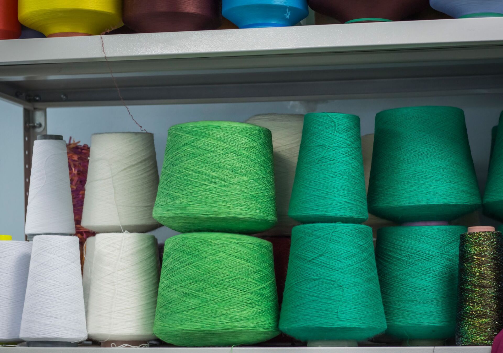 Rocchetti di filato bianco, verde brillante e verde scuro disposti su scaffali metallici.