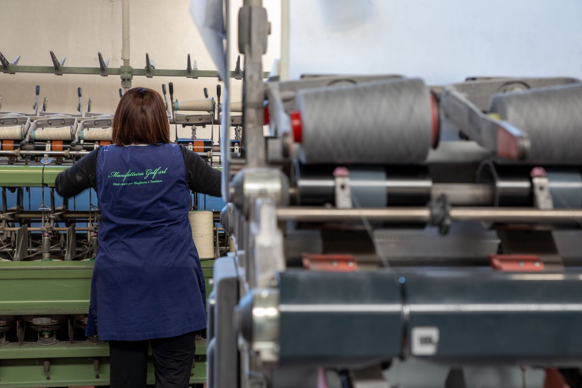 In un ambiente industriale, una persona in uniforme blu aziona macchinari tessili con bobine di filo grigie.