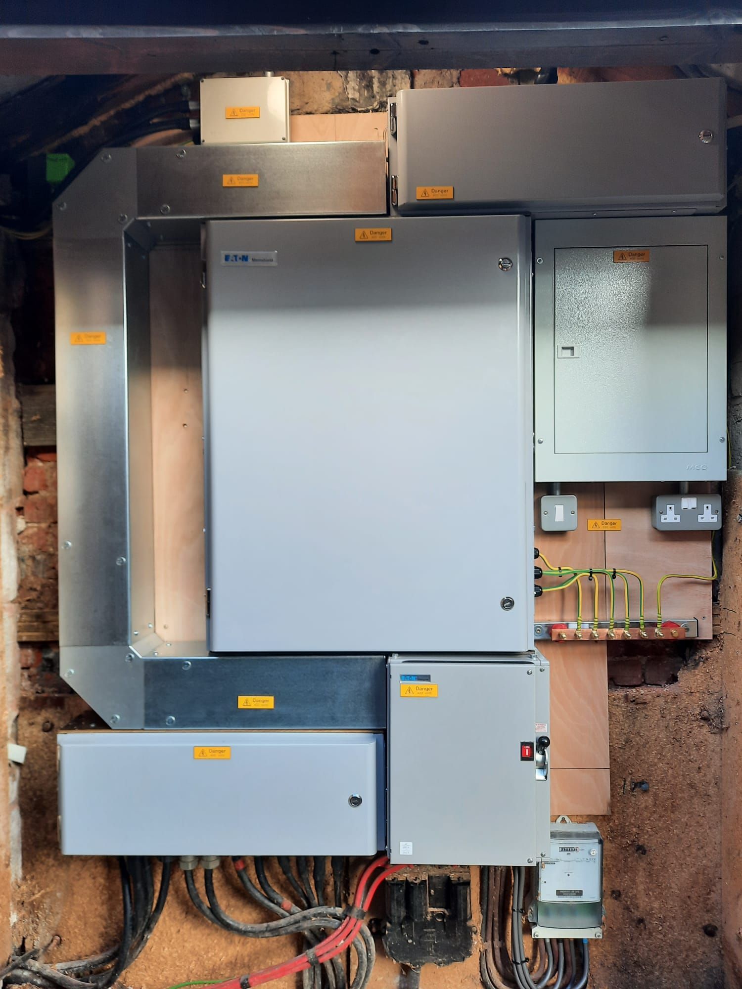 Electrical equipment mounted on a wooden surface and brick wall, including multiple gray metal boxes.