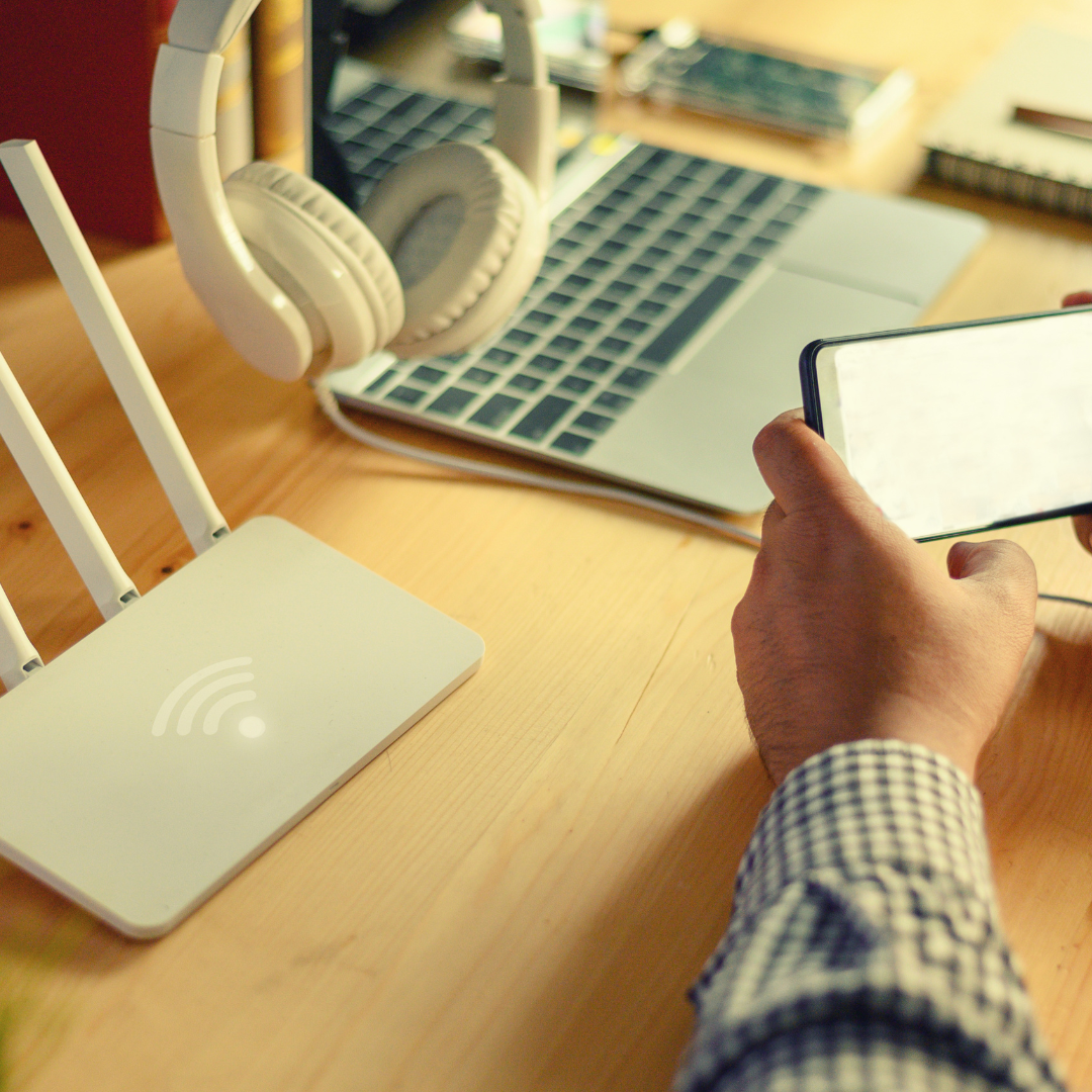 White router, laptop, headphones, and smartphone on a wooden desk. A person holds the phone.