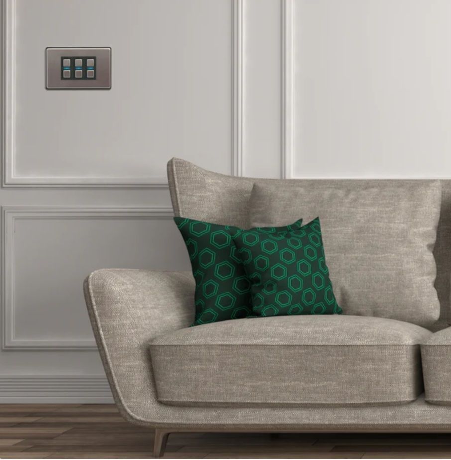Beige sofa with green patterned pillows against a white wall with light switches.