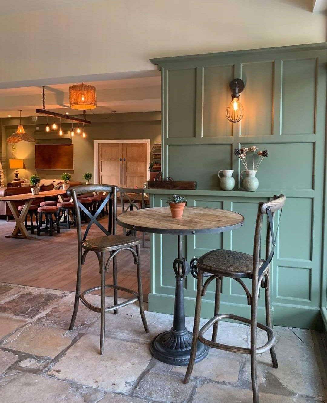 Two barstools at a small wooden table in a pub, set against a sage green panelled wall.