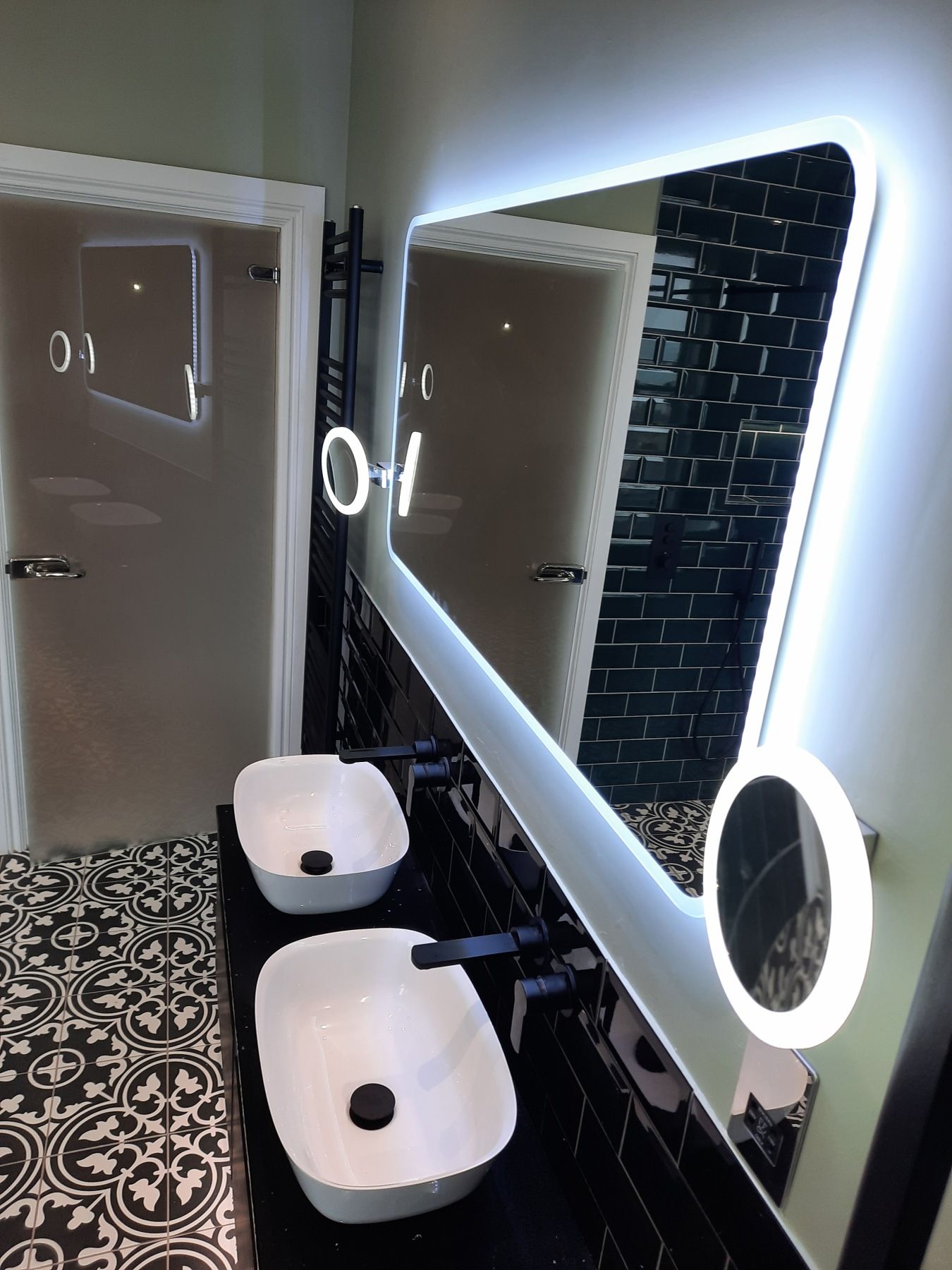 Bathroom with two white sinks, black fixtures, and a large illuminated mirror. Black tile backsplash, patterned floor.