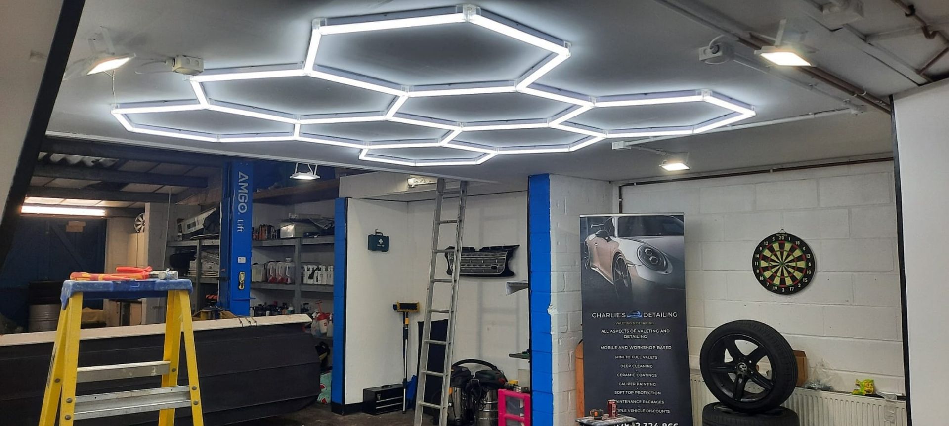 A garage interior featuring a geometric LED hexagonal lighting grid on the ceiling, a dartboard, and a tire.
