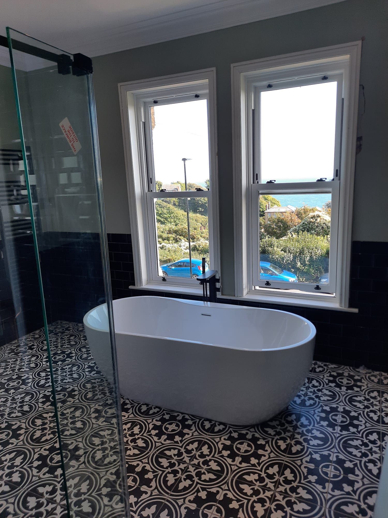 Bathroom with freestanding tub, patterned floor, and two windows overlooking a coastal view.
