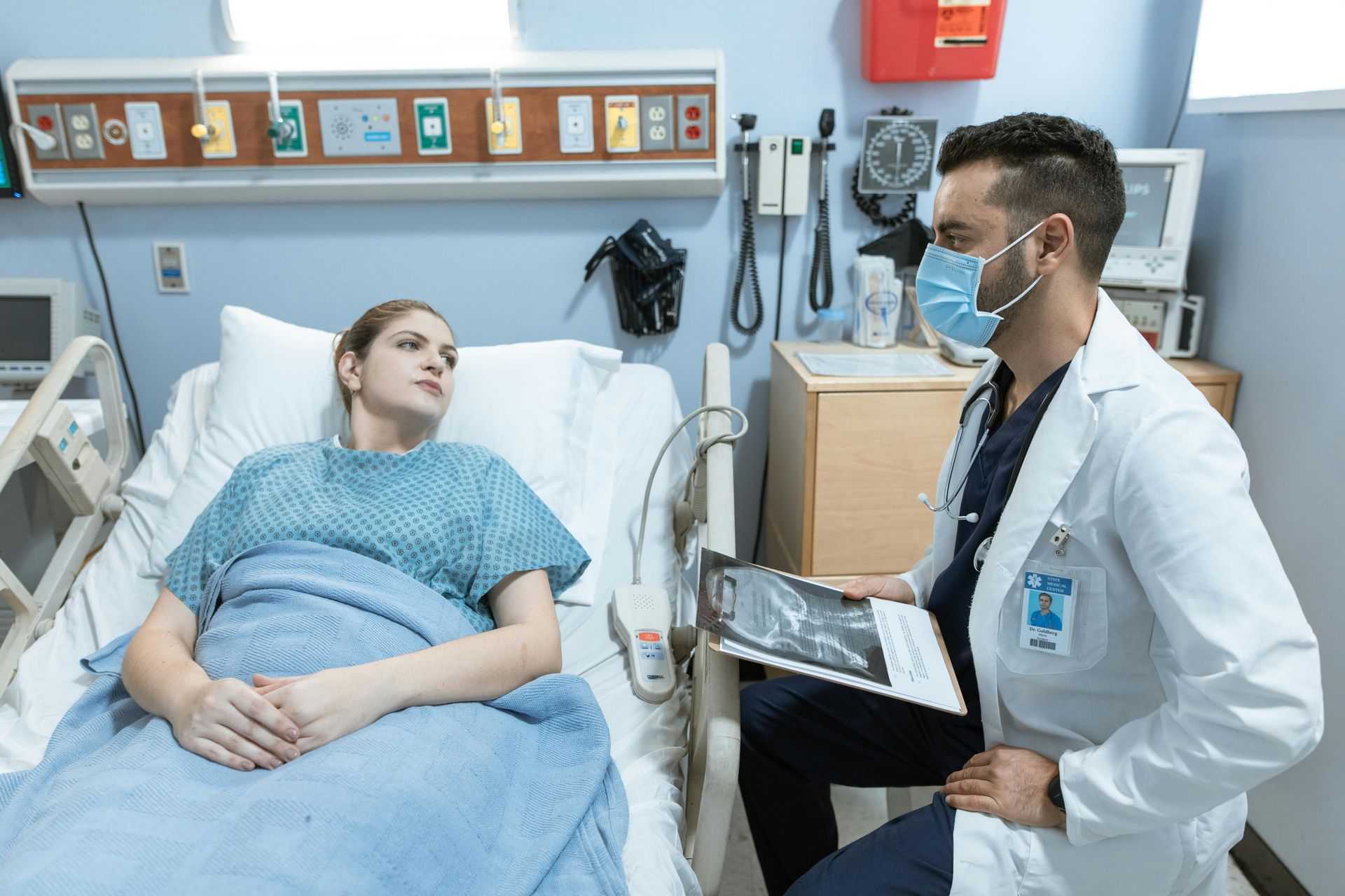 Patient in hospital bed speaking with doctor holding a clipboard in a bright hospital room