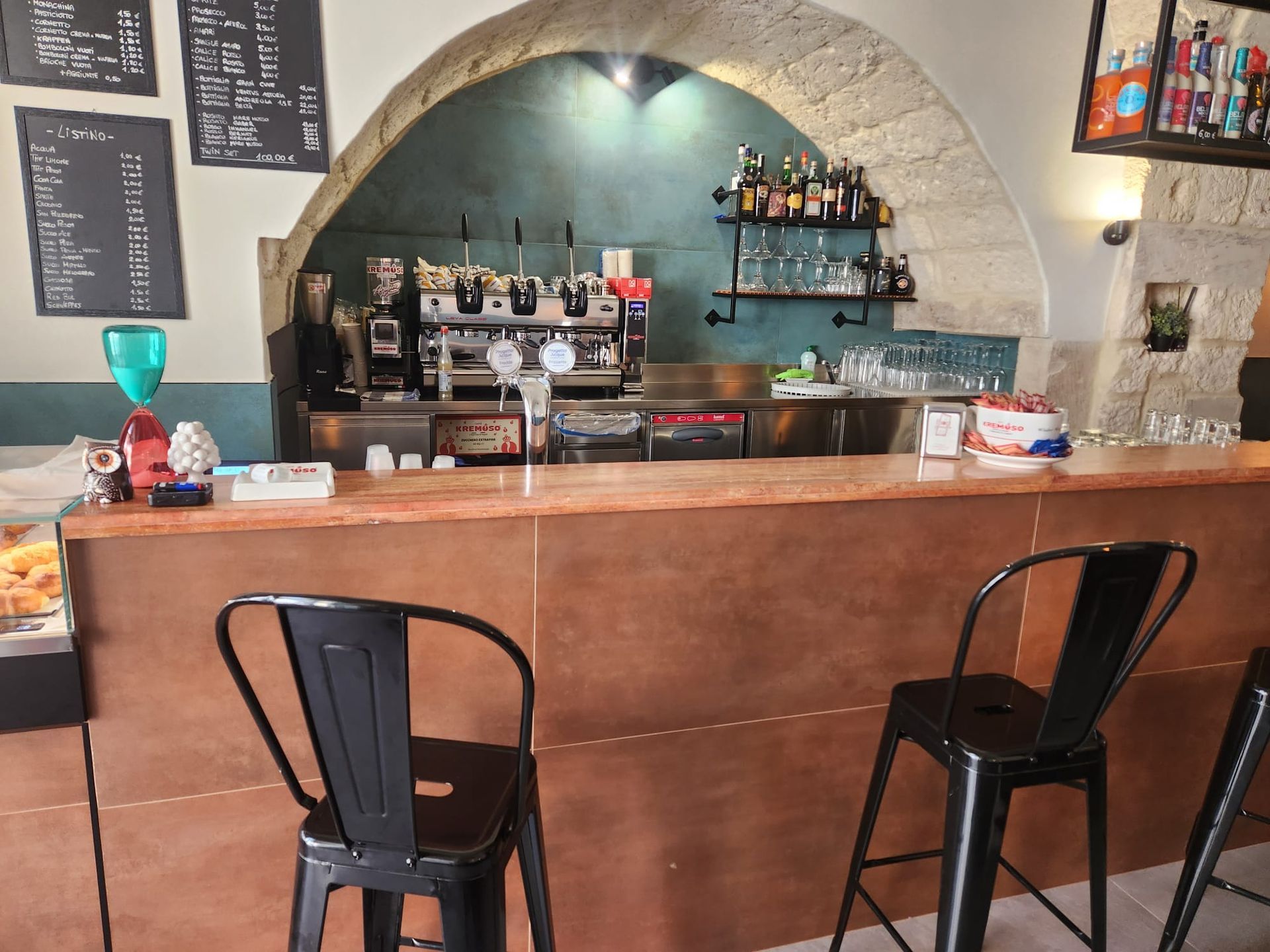 Bar rustico con bancone in legno, sgabelli neri, nicchia ad arco in pietra e scaffali di bottiglie dietro il bancone.