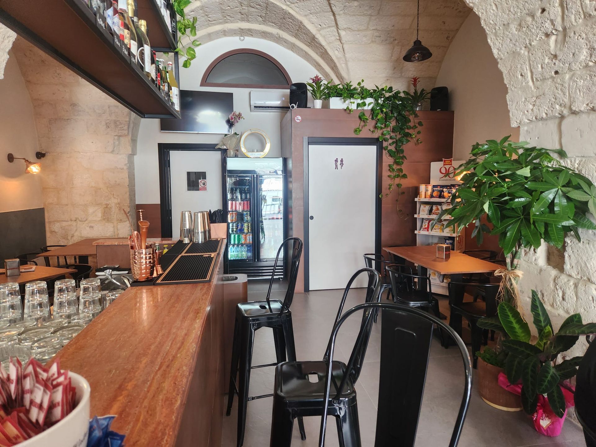 Accogliente interno di un caffè con bancone in legno, sedie nere, piante e pareti in pietra ad arco.