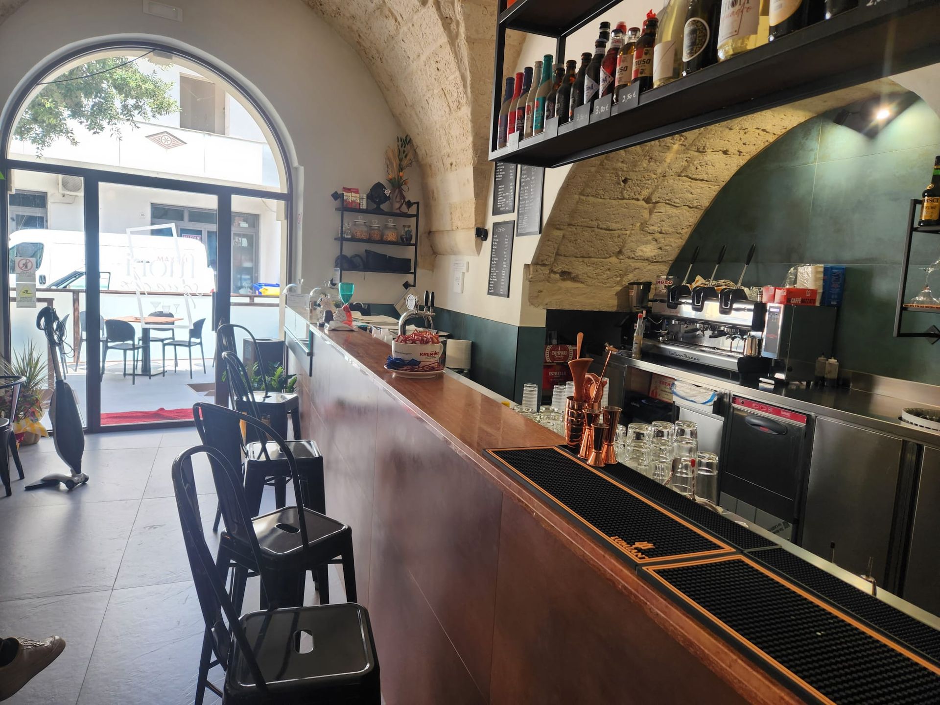 Interno vuoto di un bar-caffetteria con bancone in legno, sgabelli, porta ad arco e scaffali con bottiglie e bicchieri.