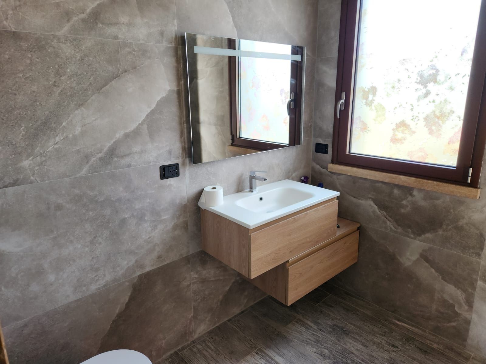 Moderno lavabo da bagno con specchio, mobiletto in legno e finestra accanto a una parete grigia con finitura materica.