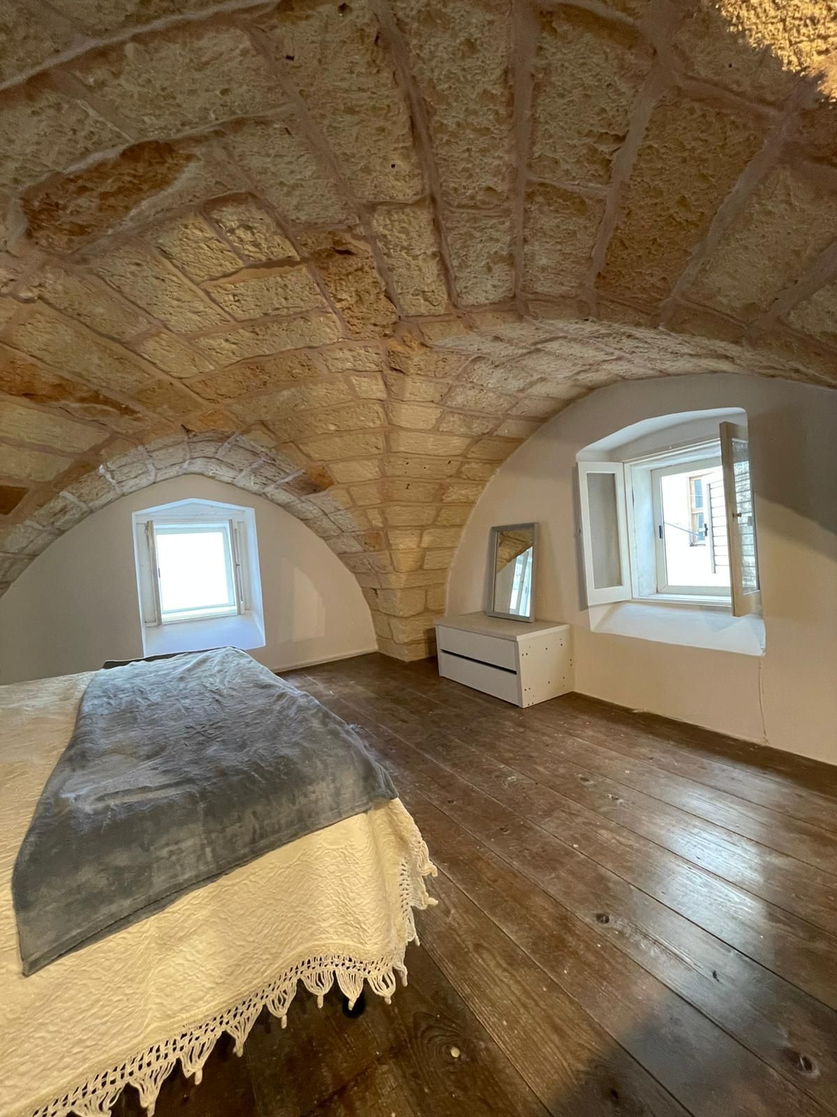 Camera da letto con soffitto a volta in pietra, pavimento in legno, letto e finestre luminose in una rustica mansarda.