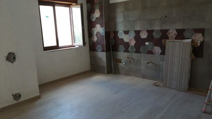 Stanza vuota e non rifinita con parete piastrellata, finestra e pavimento in legno.