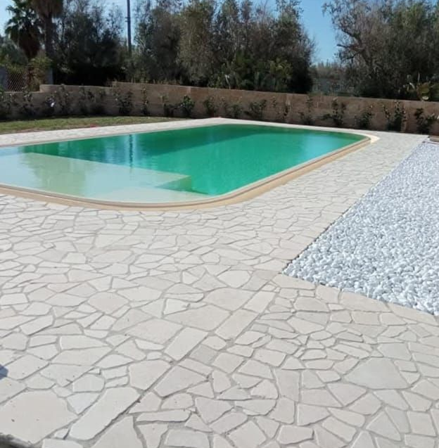 Piscina esterna turchese con pavimentazione in pietra e bordo in ghiaia in un giardino paesaggistico.