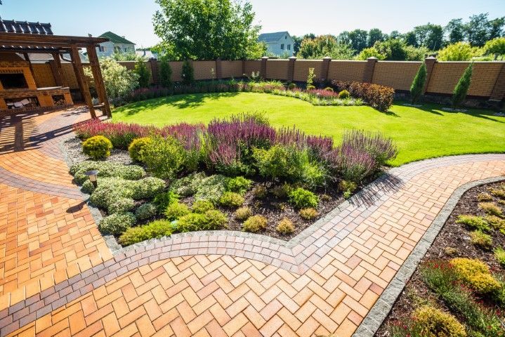A landscaped backyard with brick pavers, a wooden pergola, diverse garden plants, and a manicured green lawn.