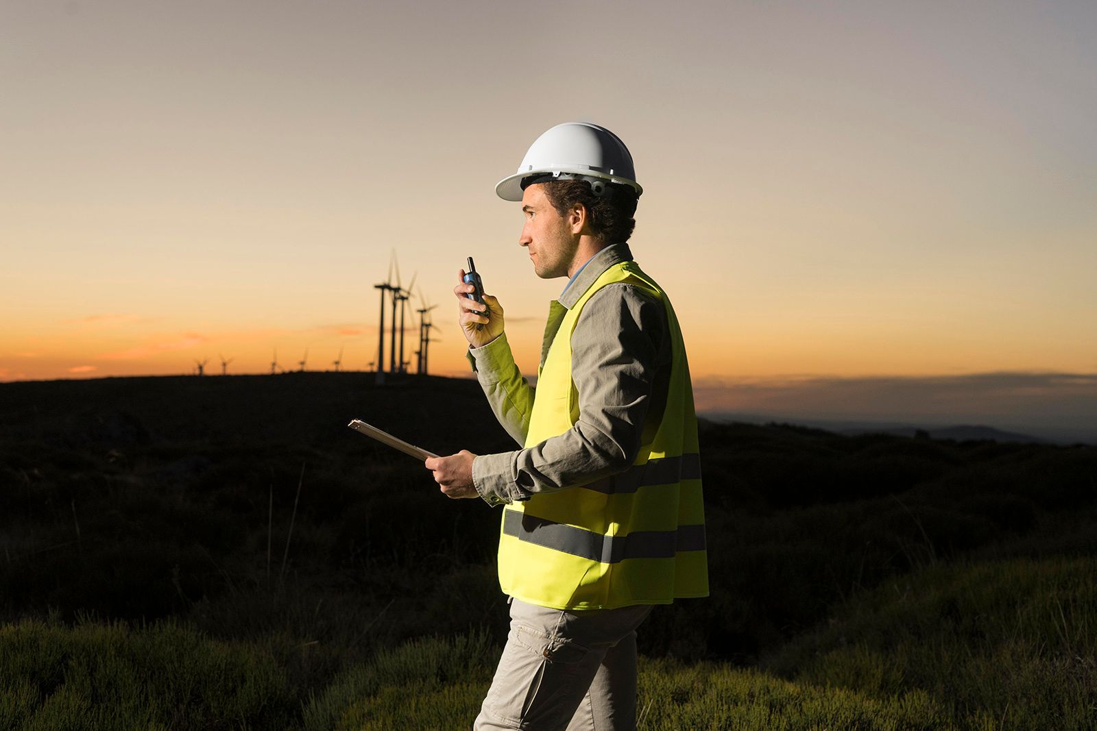 Ingeniero con casco y chaleco de seguridad, sosteniendo una radio y una tableta, con turbinas eólicas al atardecer.