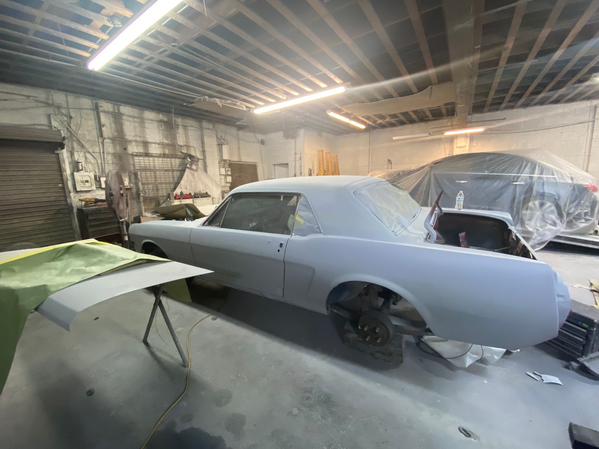 Classic car under restoration in a garage, with primer-coated body, black wheel, and covered parts nearby.