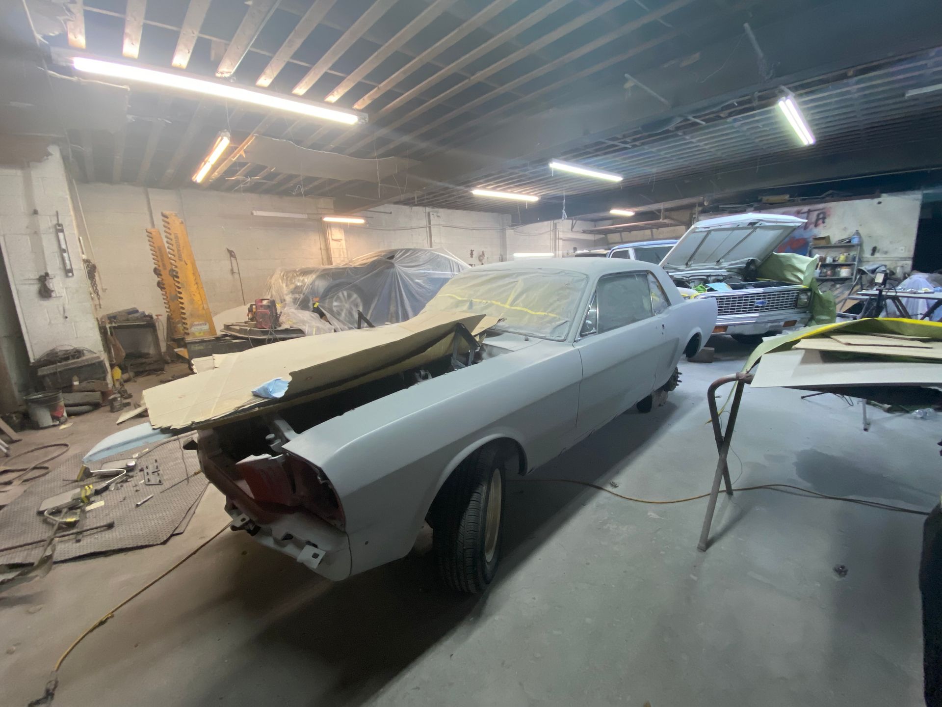 Classic car body in a cluttered auto repair shop, primed gray with open hood and trunk.