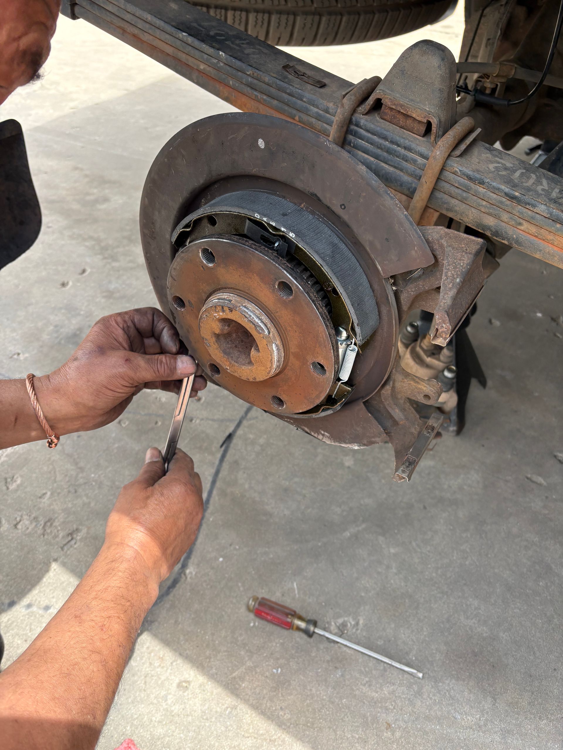Person working on a rusty vehicle brake assembly with hand tools on a garage floor.