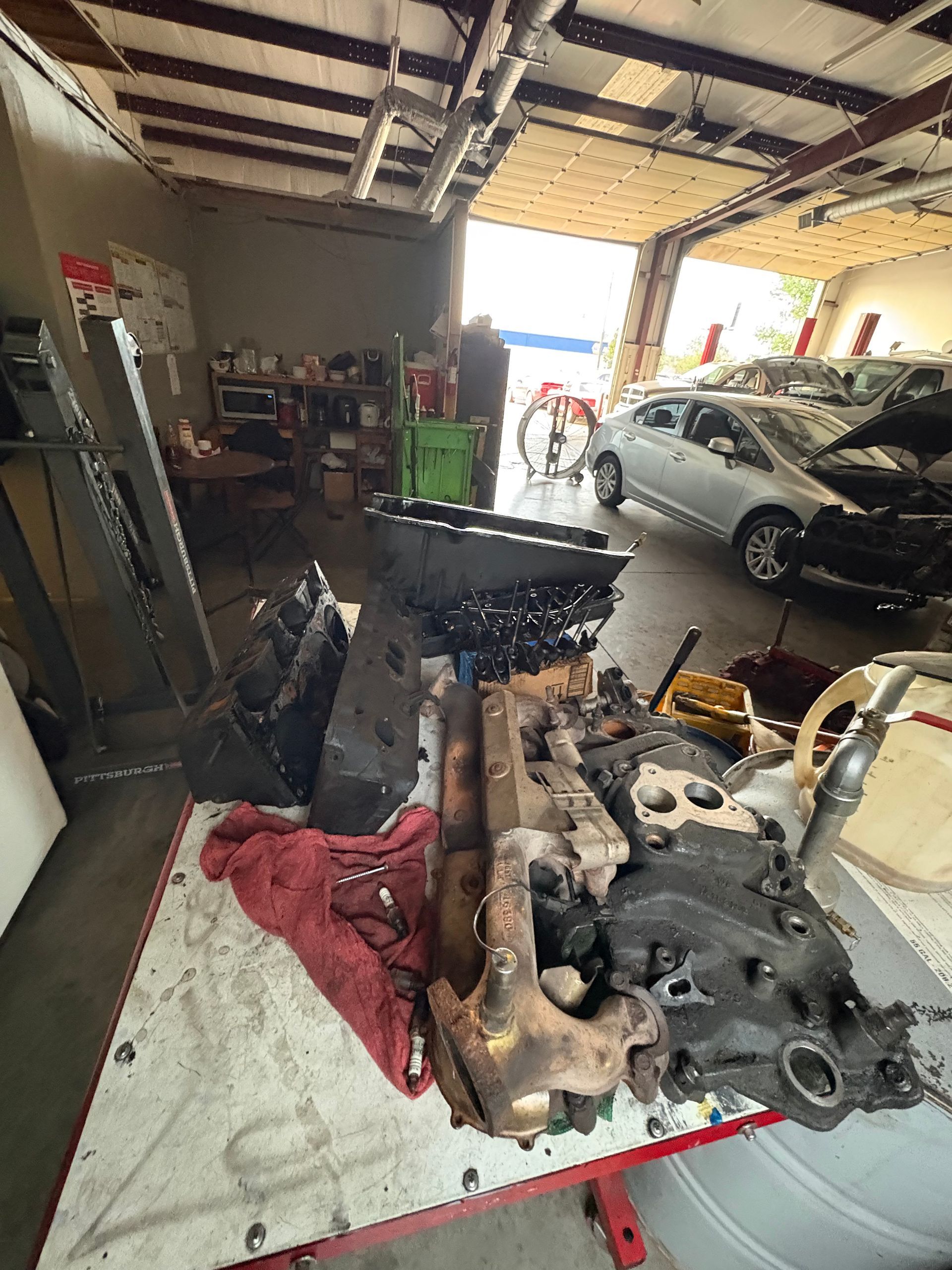 Car repair shop with engine parts on a workbench and a silver car parked inside.