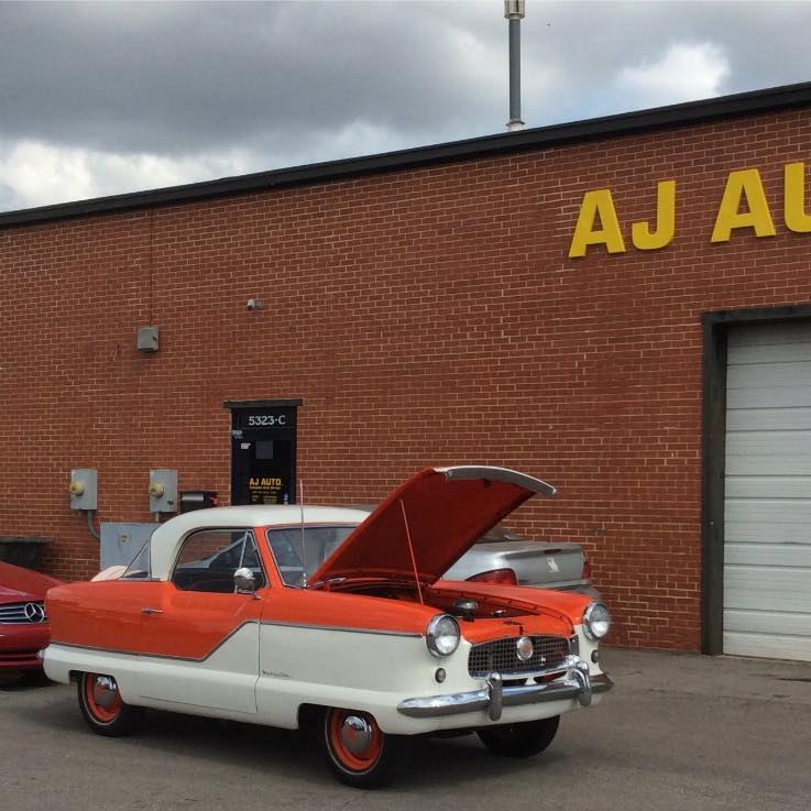 Orange-and-white vintage car with hood open parked outside a brick auto shop