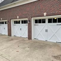 Three white carriage-style garage doors with black hardware and windows on a red brick residential building.