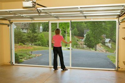 A woman standing inside a garage looking out through a white-framed retractable screen door onto a paved driveway