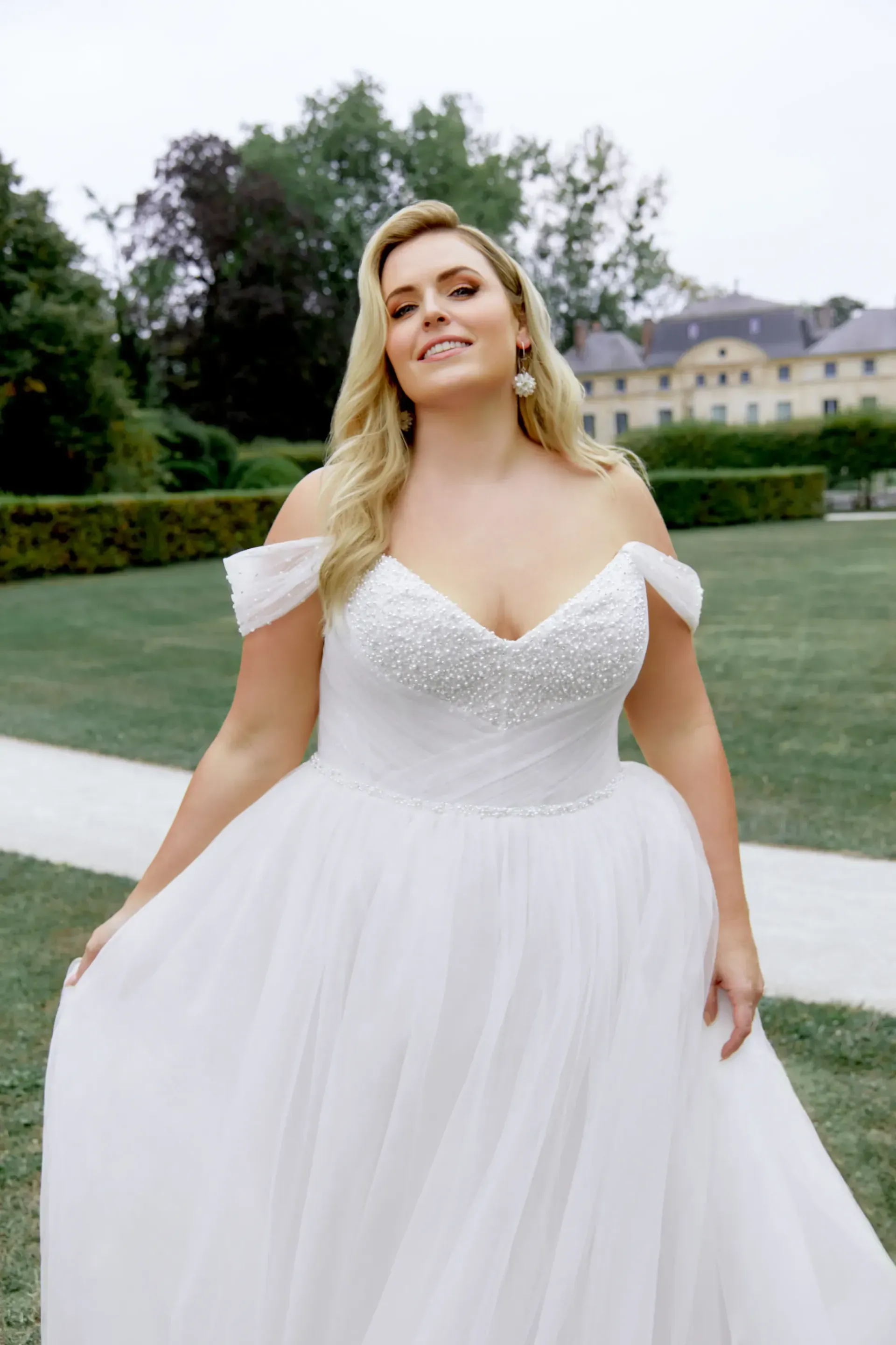 Woman in white off-the-shoulder wedding dress smiles outdoors. Ballgown, tulle skirt.