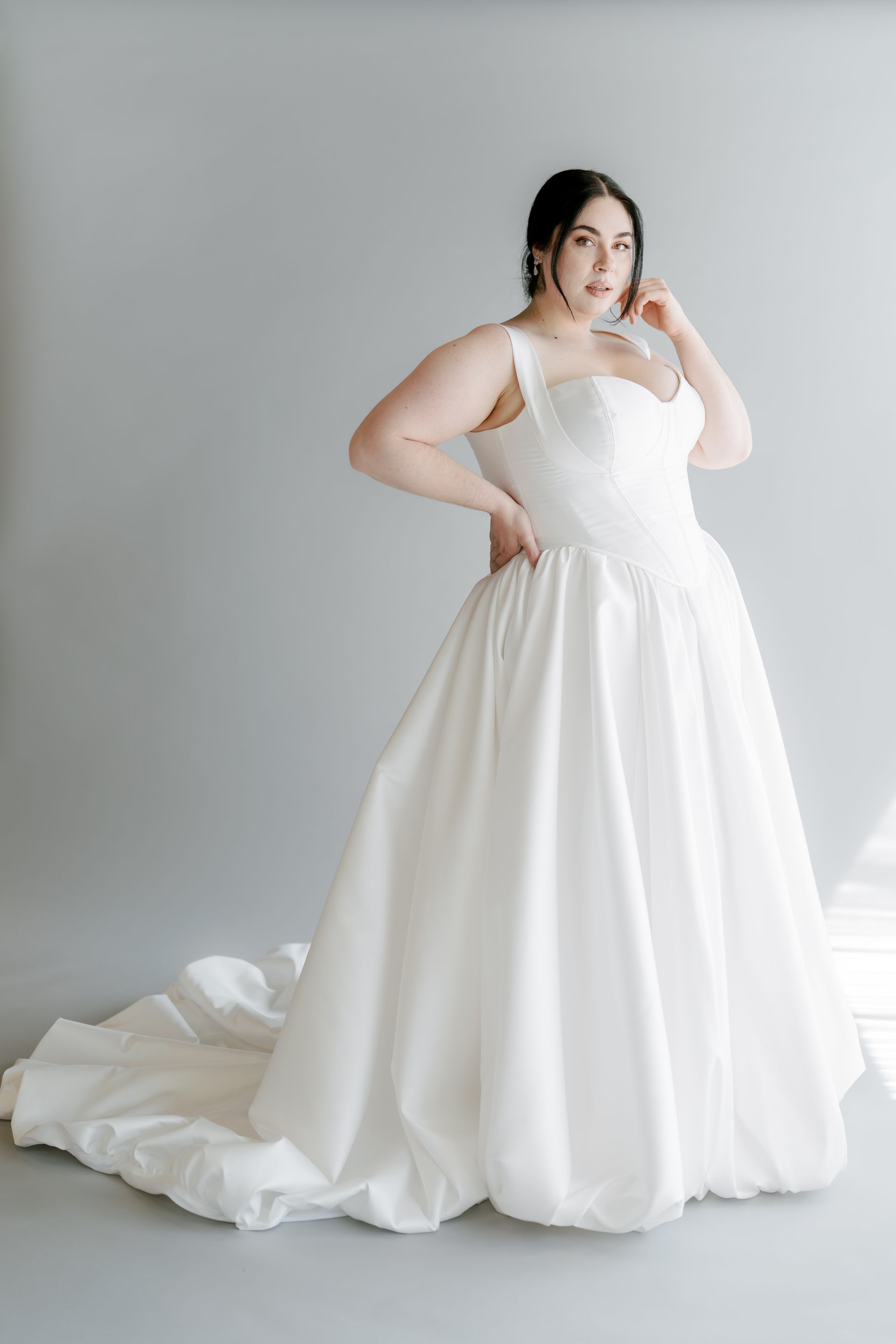 Woman in a white wedding gown with a full skirt and train, posing with hand on hip.