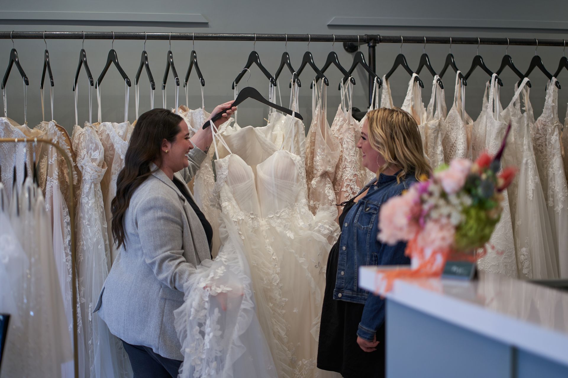 Woman assisting another woman selecting a wedding dress from a rack in a bridal shop.