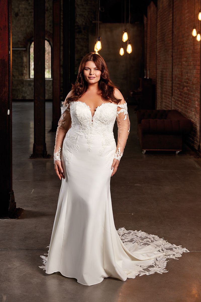 Woman in white off-shoulder wedding gown, long sleeves, train, in a brick-walled industrial space.