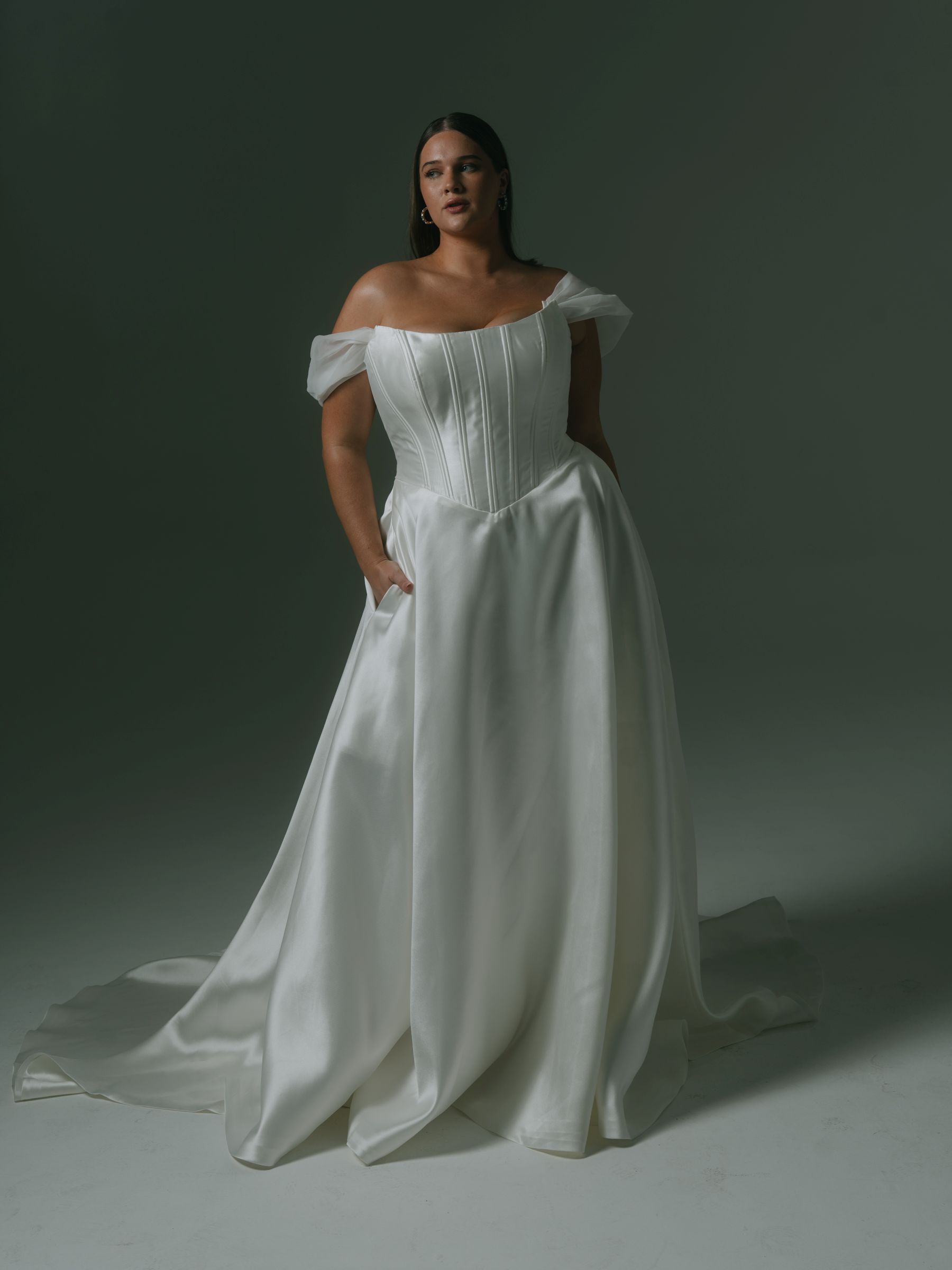 Woman in a white off-shoulder wedding gown, hands in pockets, standing in a studio with a slight train.