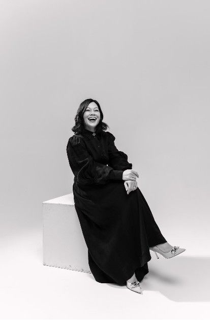 Woman seated, smiling, in black dress, heels, on white cube, studio background.