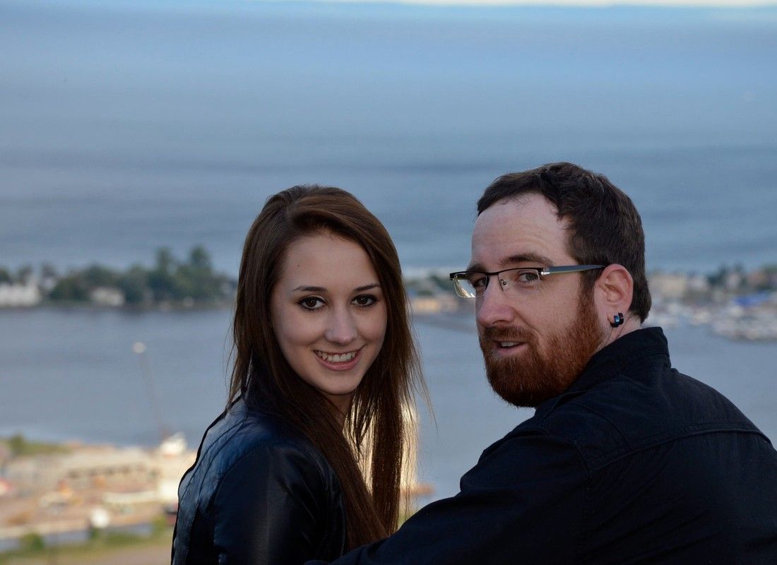 Two people smiling by the water at sunset, with a blurred shoreline in the background