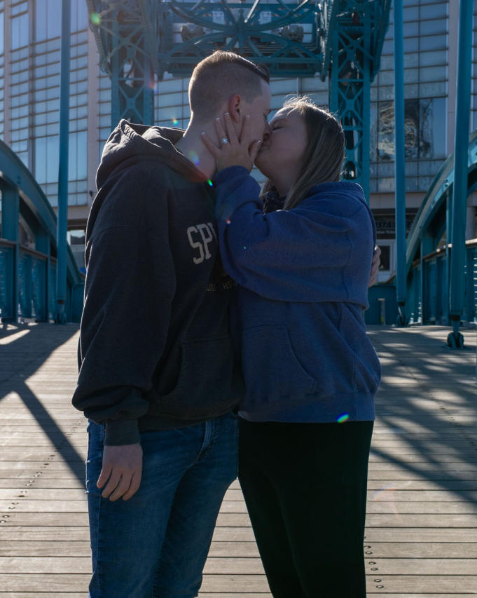 Two people kissing outdoors by a blue metal bridge in sunlight