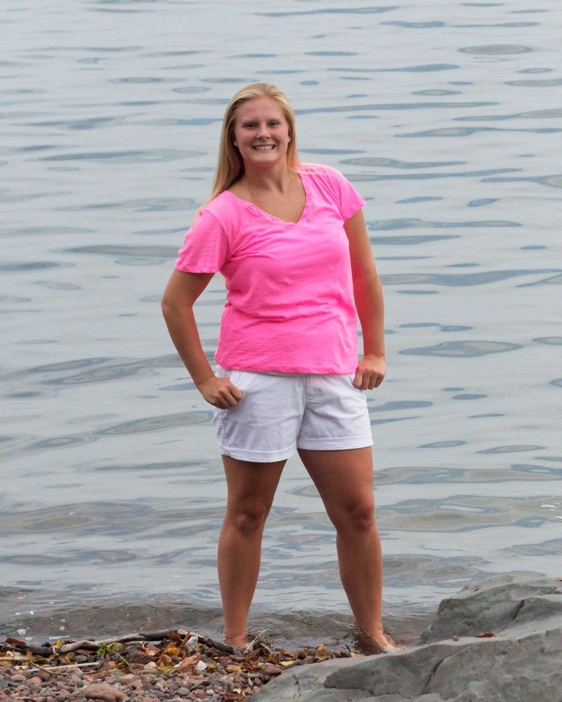 Woman in a pink shirt and white shorts standing on a rocky lakeshore, smiling at the camera.