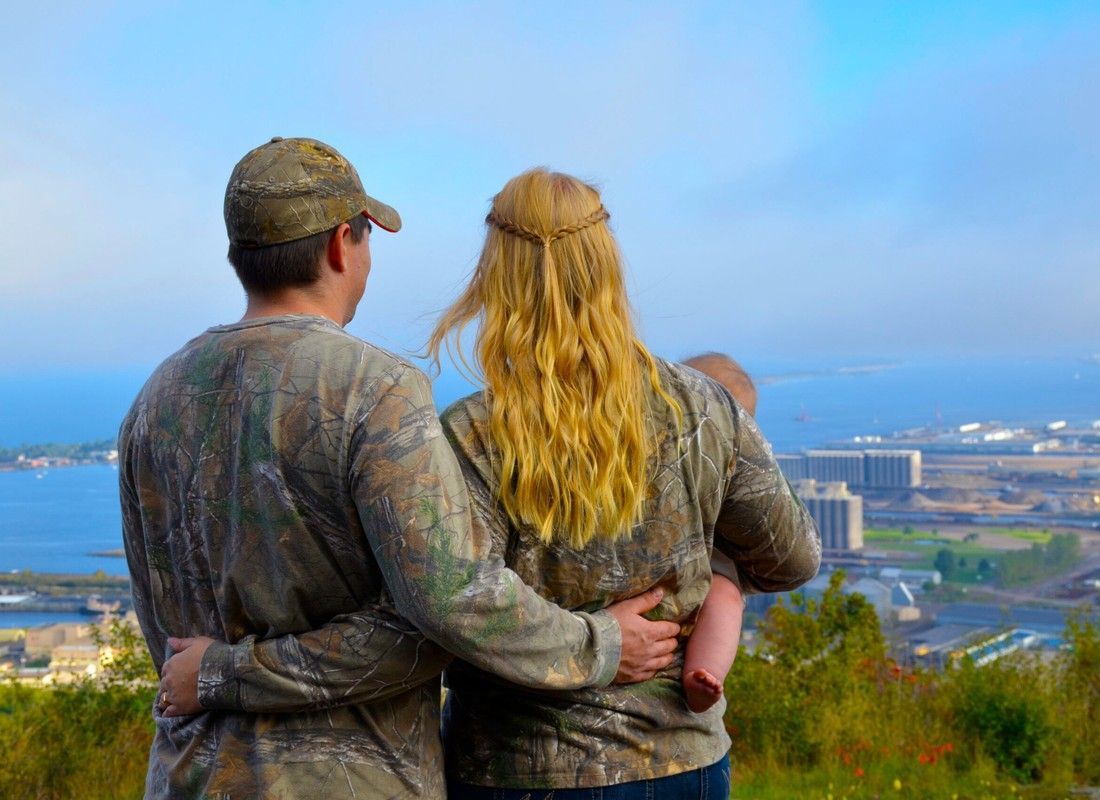 Two people in camouflage jackets stand embracing on a hill overlooking a coastal city and sea