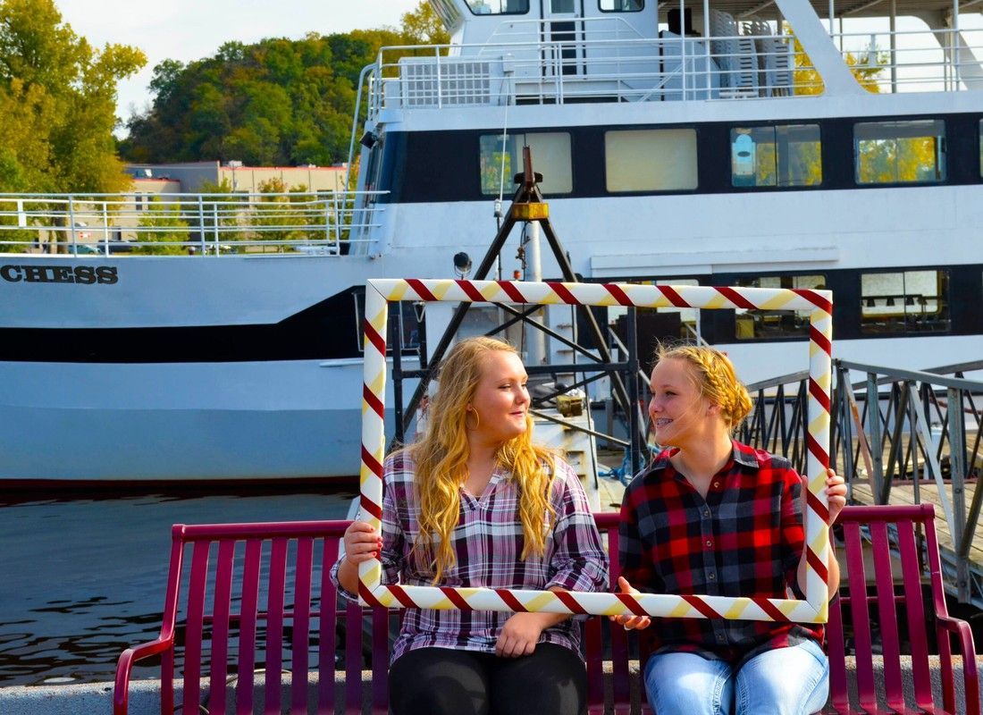 Two people sitting on a red bench by a docked boat, chatting outdoors in plaid shirts.