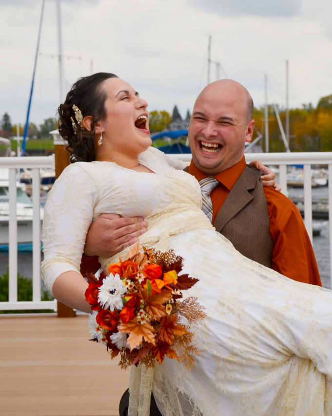 Laughing couple in wedding attire, groom carrying bride with autumn bouquet on a dock.
