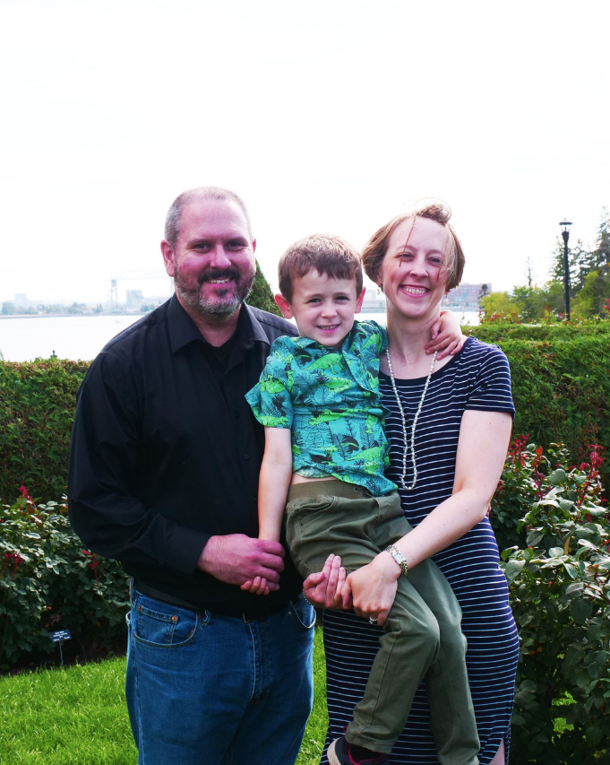 Three people posing outdoors in a garden, smiling with flowers and trees behind them.