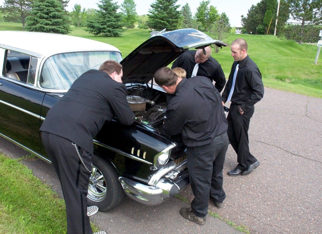 Four men inspect a black vintage car with the hood open on a grassy roadside.