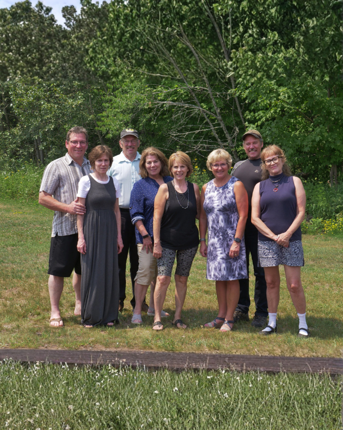 Seven people pose together outdoors in front of green trees and grass, dressed casually in summer clothes.
