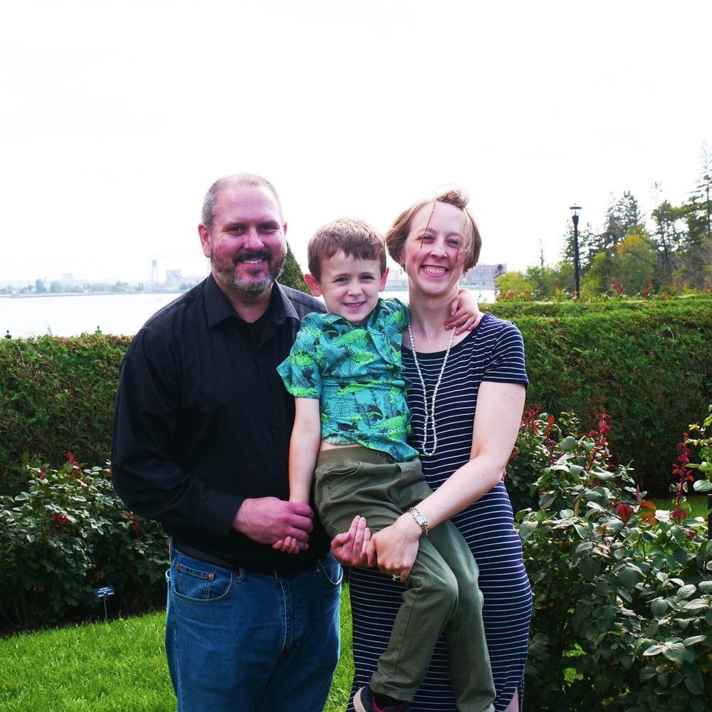 Three people posing in a garden, smiling, with flowers and trees behind them.