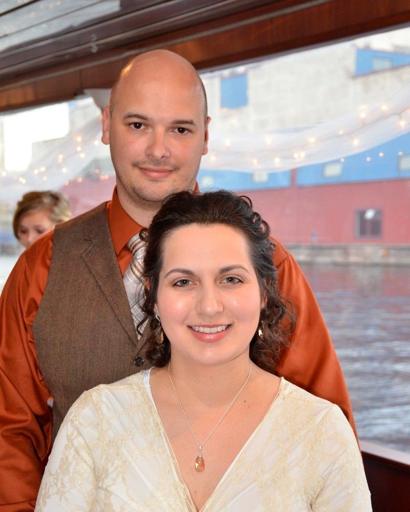Two people smiling indoors by a window, with the woman in front and the man standing behind her.