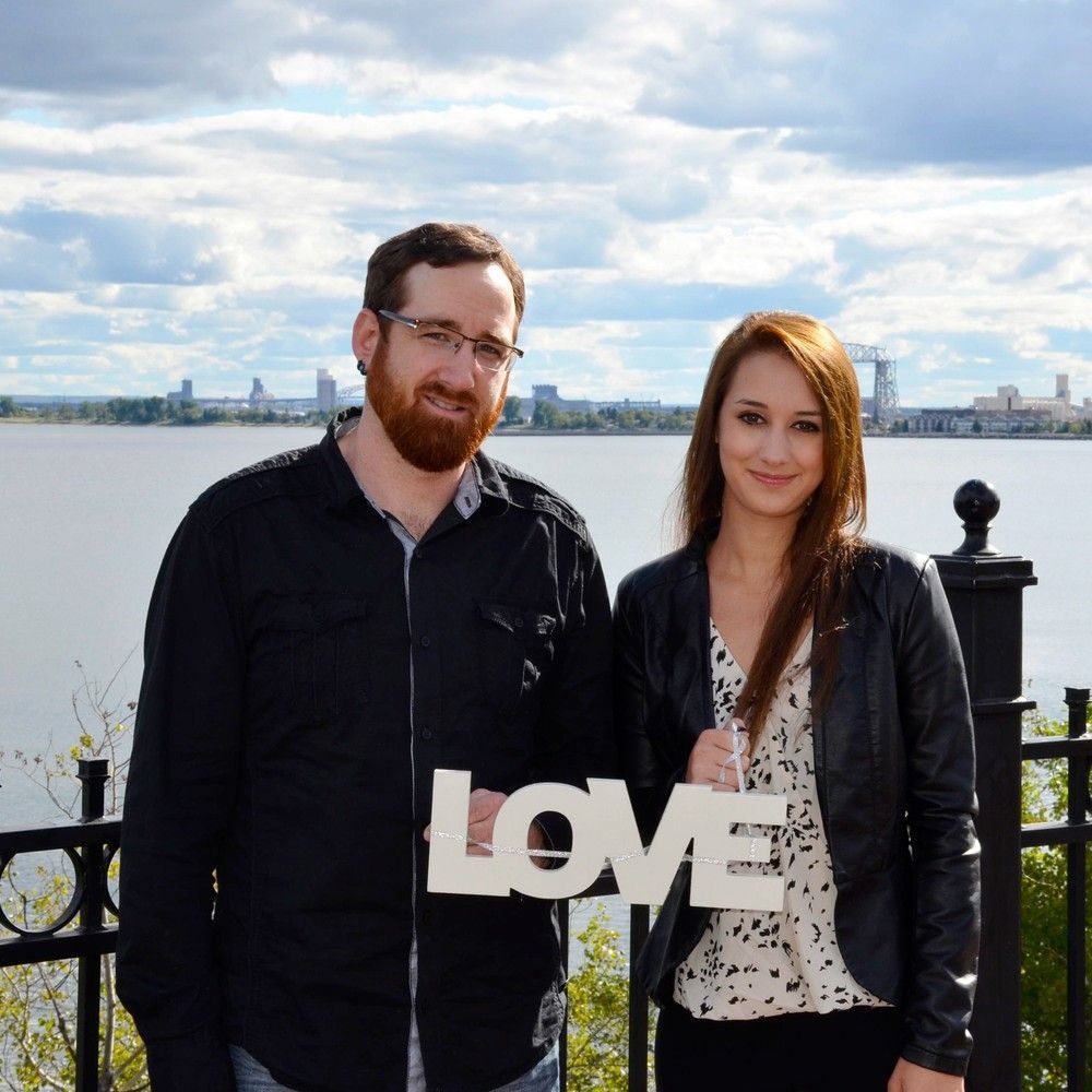 Two people standing by the water holding a white “LOVE” sign, with a city skyline in the background.