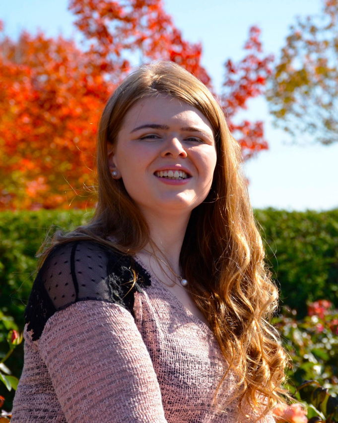 Smiling woman outdoors with long brown hair, wearing a pink sweater, with autumn trees in the background