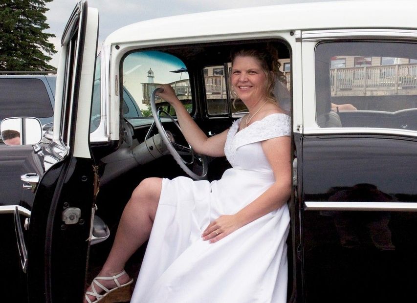 Woman in a white dress sitting in the open doorway of a vintage car, smiling.
