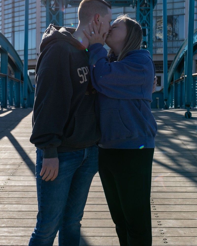 Two people kissing on a sunlit bridge, wearing dark hoodies and jeans.