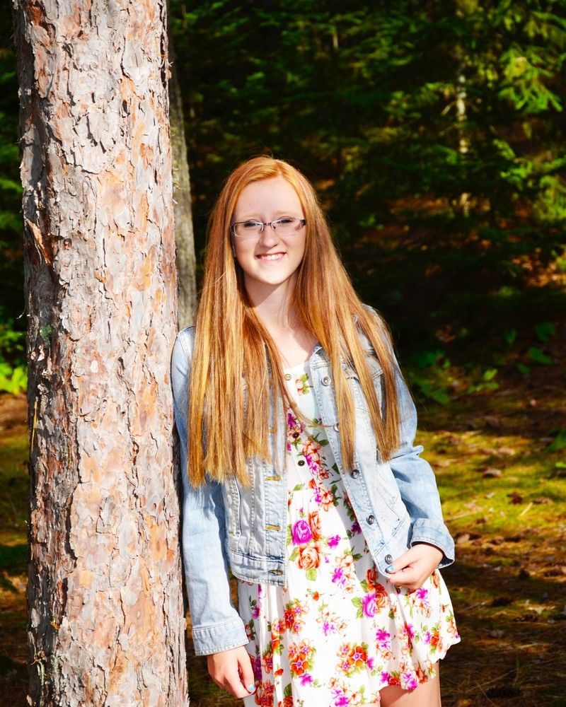 Girl with long red hair in a floral dress and denim jacket, standing by a tree outdoors and smiling